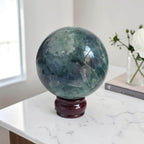 Dark green fluorite crystal sphere on glossy dark pedestal stand, bright highlights on face