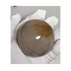 Honey calcite crystal sphere held in white gloves, translucent beige with warm amber patch