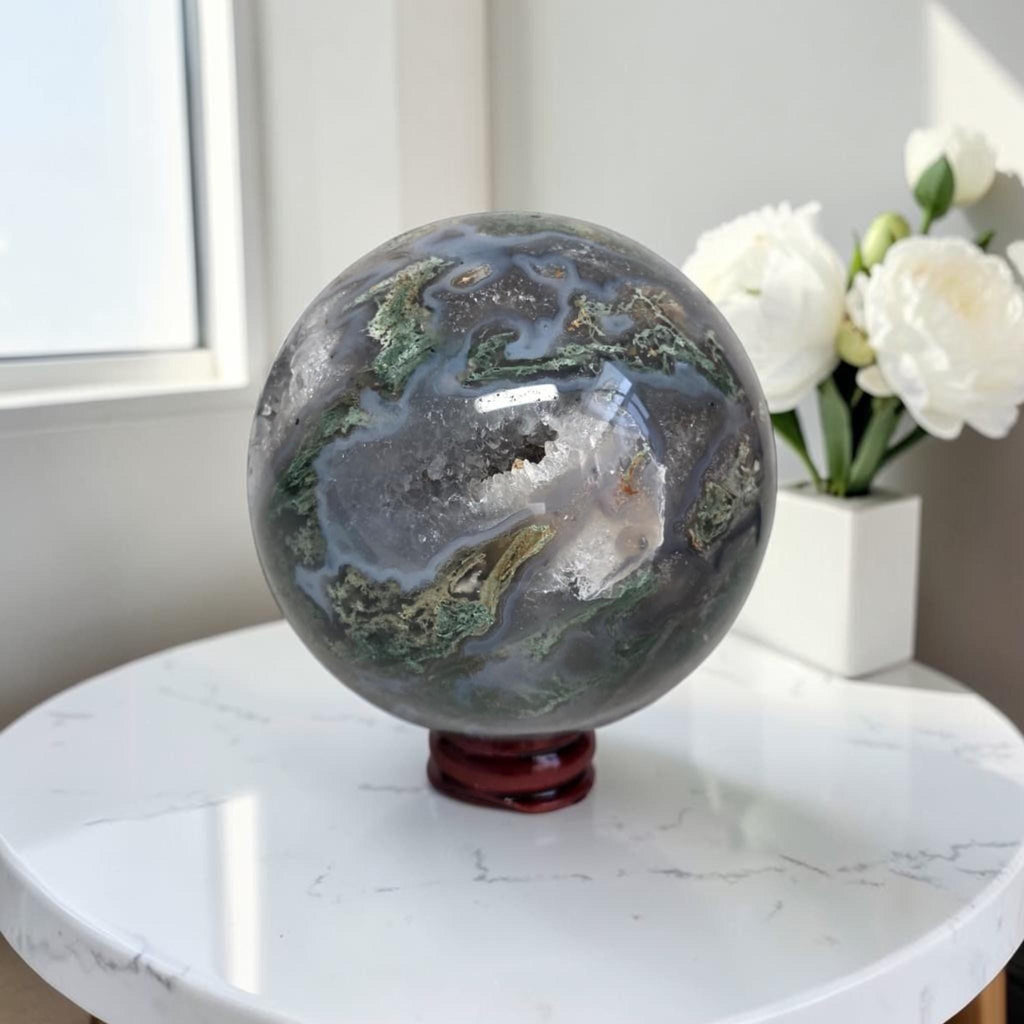 Moss agate geode sphere on round base: dark gray-blue with green patches and clear druzy