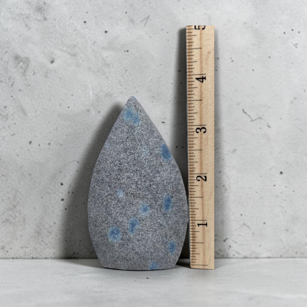 Teardrop polished stone next to measuring ruler, gray with blue spots, pointed shape