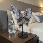 Butterfly sculpture on a stand next to a clear glass vase on a wooden table with a blurred sofa in the background.