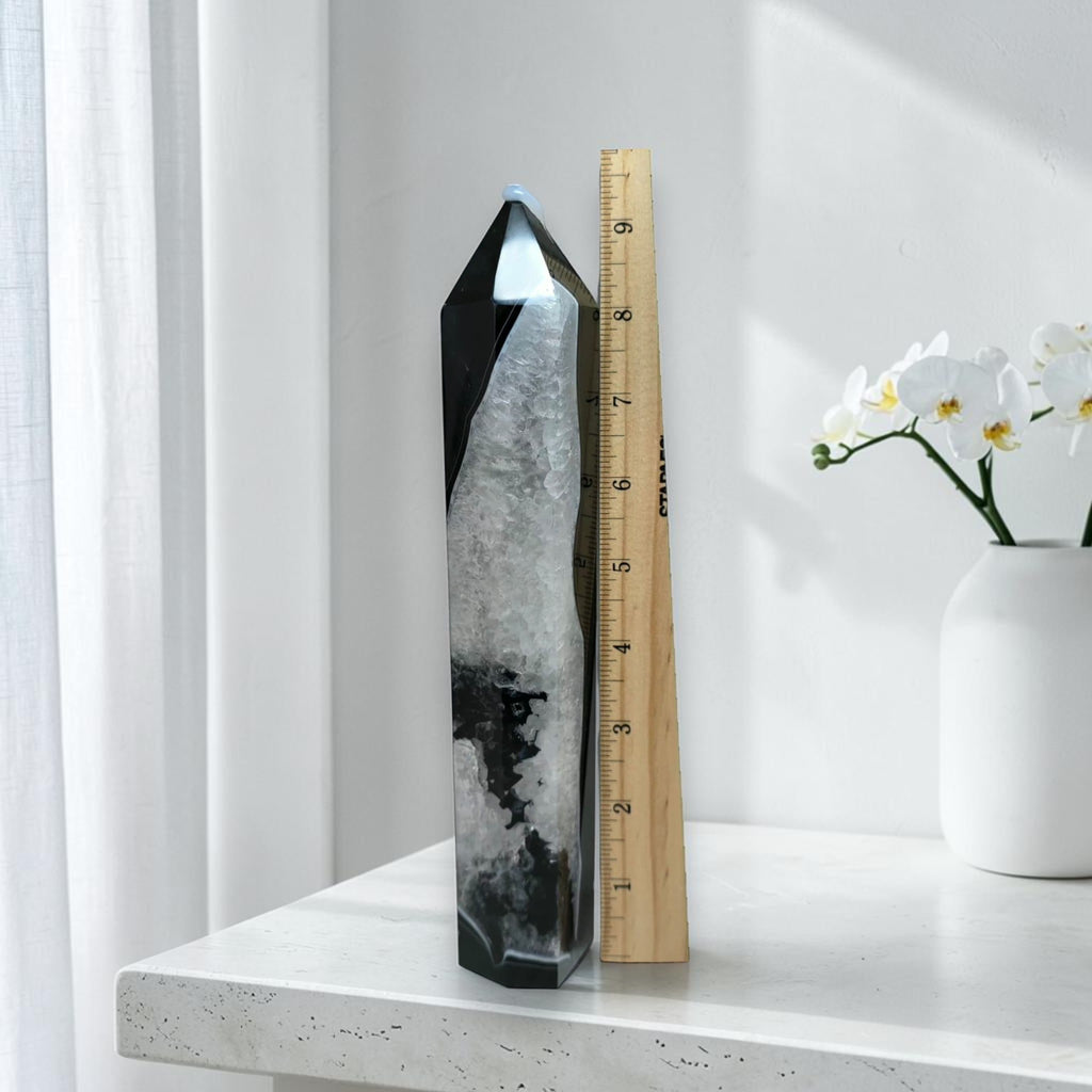 Black agate quartz tower next to a ruler showing approximately 8 inches in height, on a white marble surface with orchid flowers