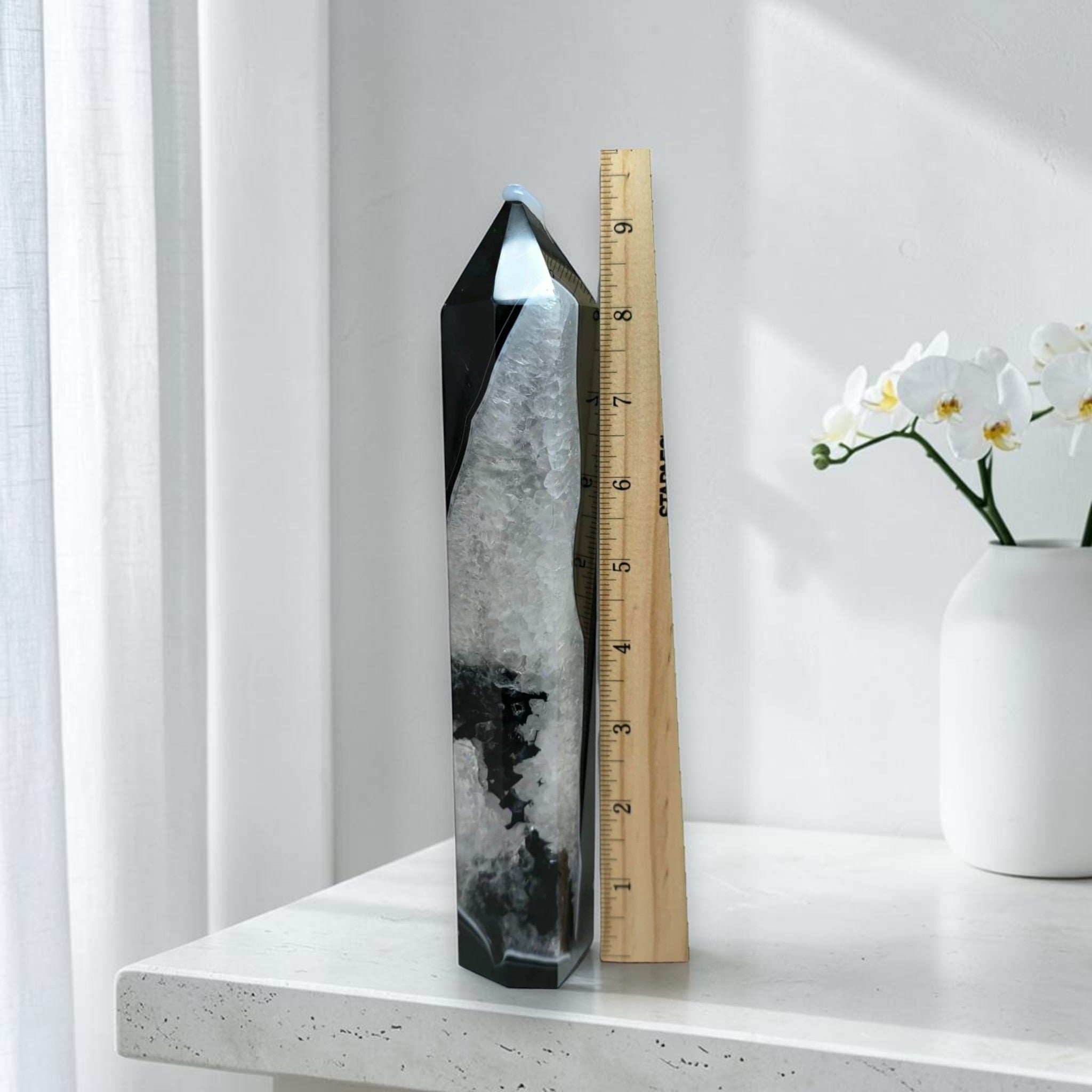Black agate quartz tower next to a ruler showing approximately 8 inches in height, on a white marble surface with orchid flowers