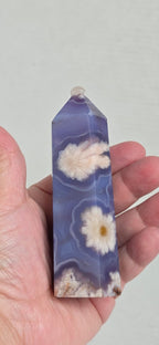 Hand holding a marbled purple and white crystal tower with floral inclusions against a plain background