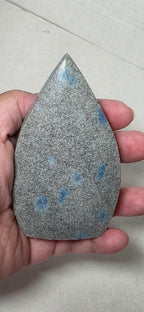 Teardrop polished stone held in hand, gray with blue dot spots and sharp tip