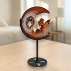 Side view of amber moon crystal carving with fairy figures and druzy geode on black stand