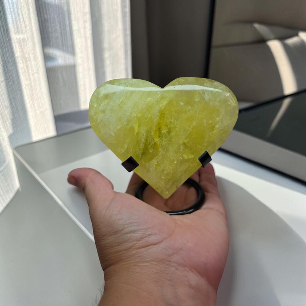 Heart-shaped yellow object held by a hand with a window and curtain in the background