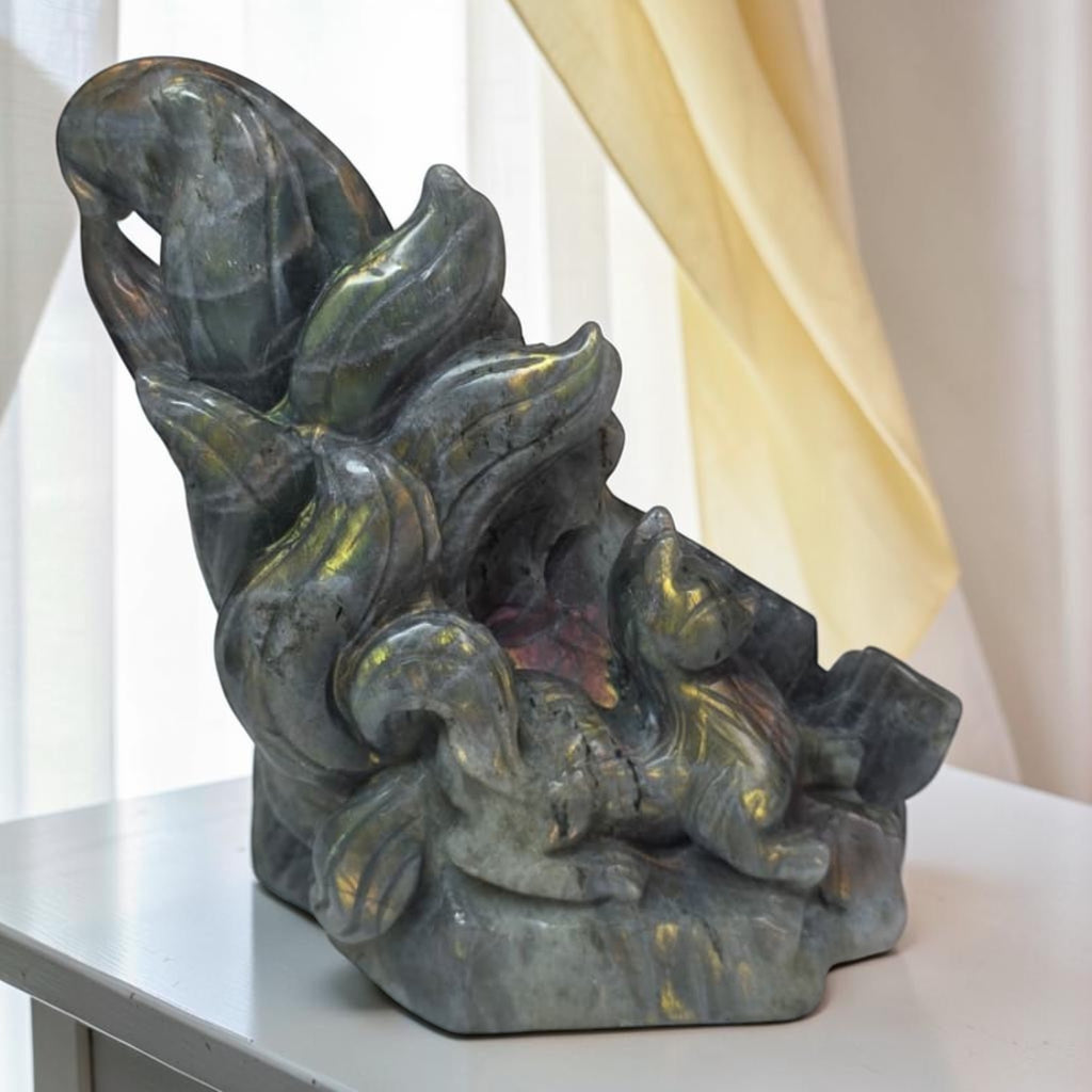 A carved stone sculpture depicting a stylized figure with flowing, leaf-like forms, displayed on a white table in front of a soft cream curtain backdrop.