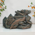 A carved dragon figurine made of natural labradorite, featuring a textured metallic sheen with green, gray, and coppery-brown tones, displayed on a white shelf against a white wall with decorative foliage in the corners.
