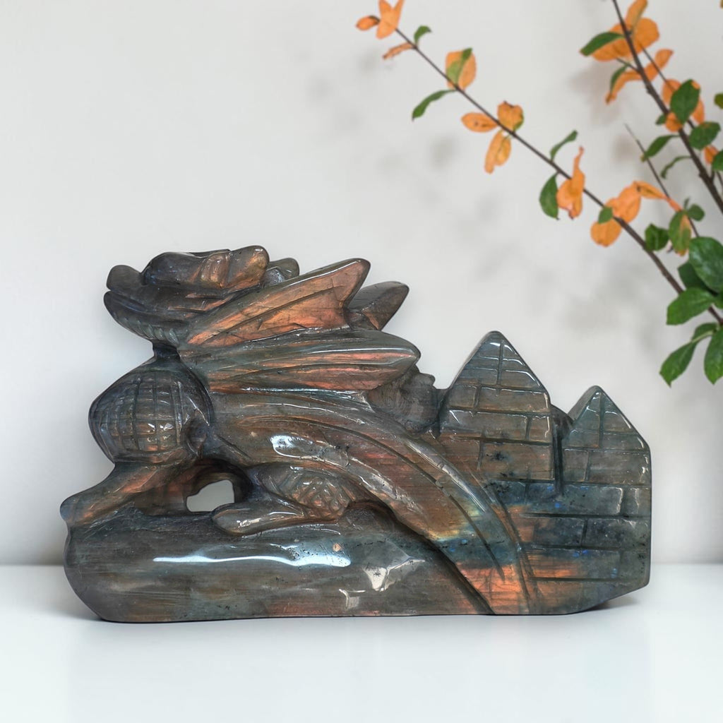 A carved dragon figurine made of iridescent labradorite stone, featuring coppery-orange and blue-green flashes, displayed on a white surface with a white wall and orange foliage in the background.