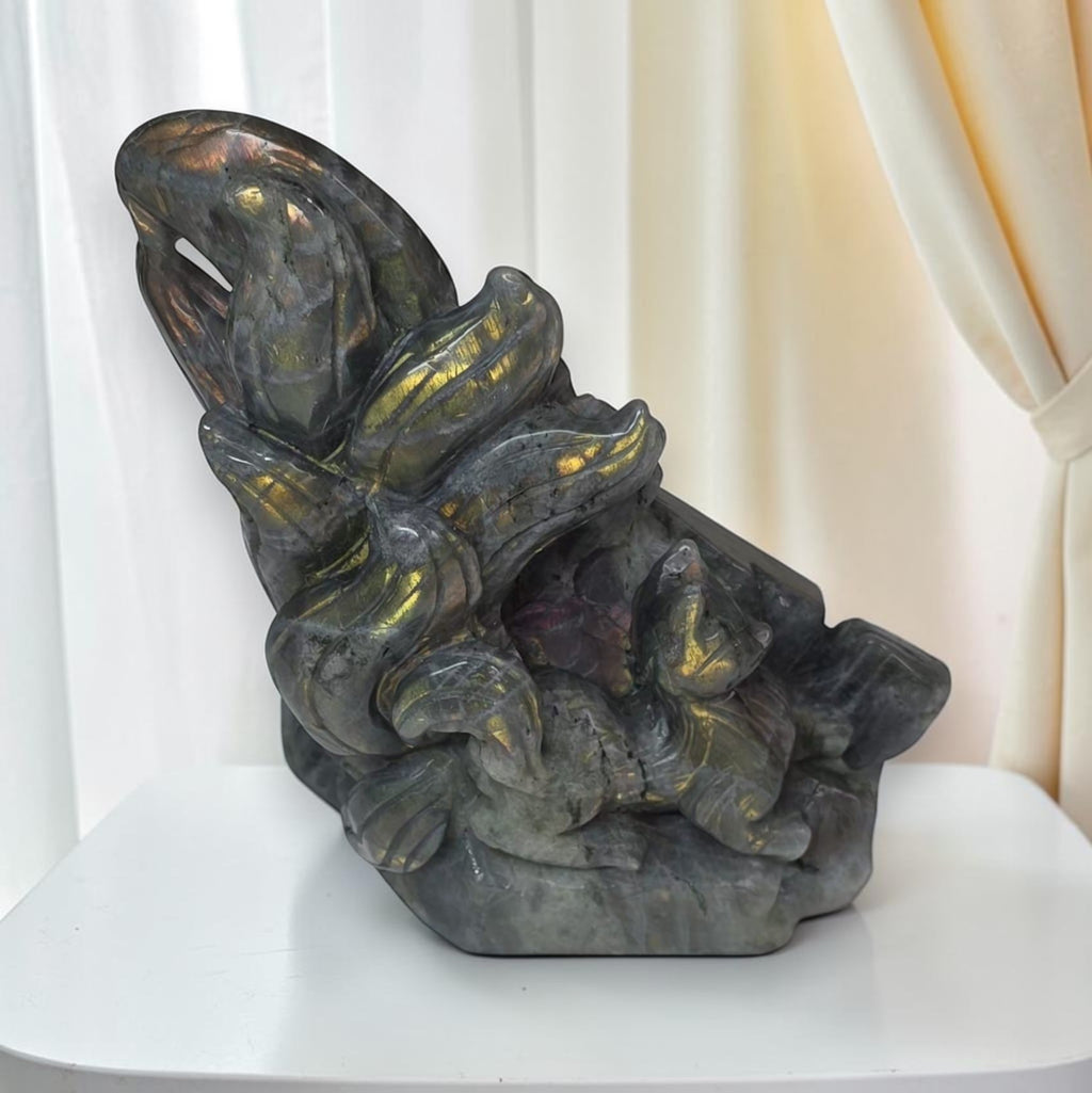 A carved stone sculpture depicting a stylized figure with flowing hair, shown on a white table against a soft cream curtain background.