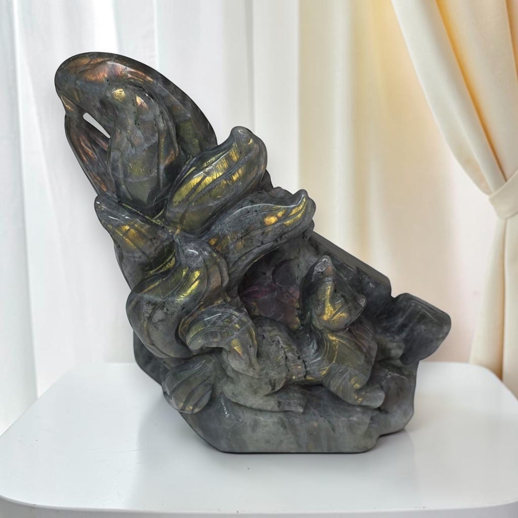 A carved stone sculpture depicting a stylized figure with flowing hair, shown on a white table against a soft cream curtain background.