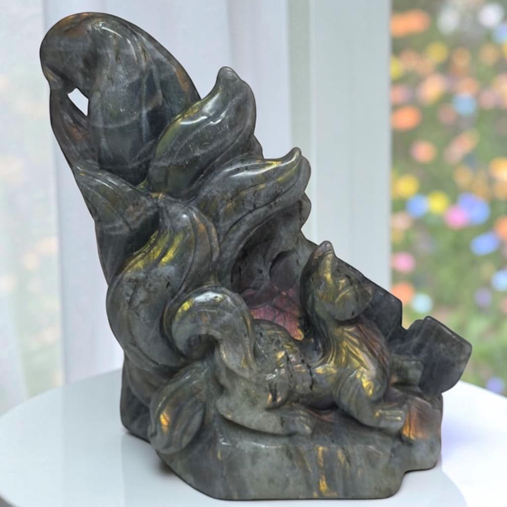 A dark gray stone carving of a fox sitting among stylized flames, displayed on a white surface with a softly blurred window background.