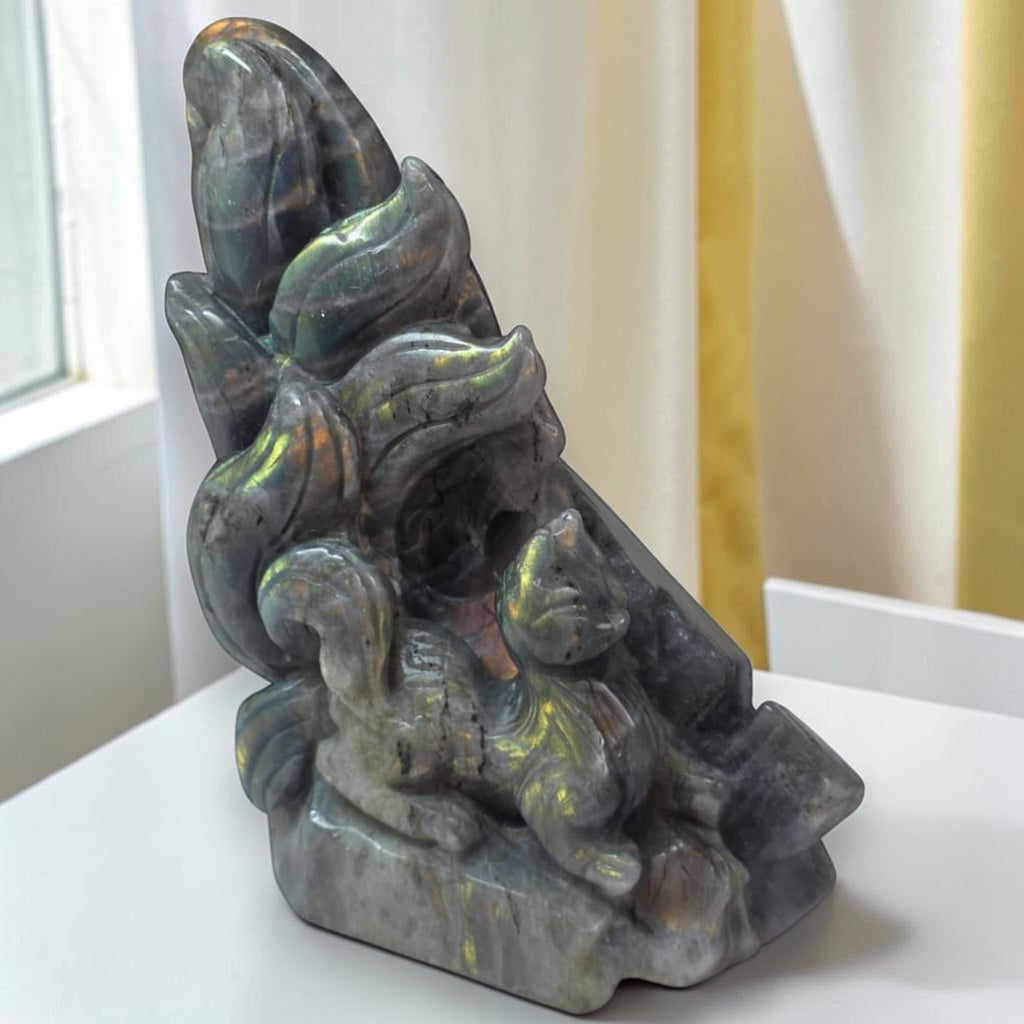 A carved stone figurine of a fox and cat on a base, displayed on a white surface with a window and curtains in the background.