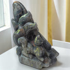 A carved stone figurine of a fox and cat on a base, displayed on a white surface with a window and curtains in the background.