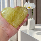 Heart-shaped yellow stone held in a hand with a white vase and orchid in the background.