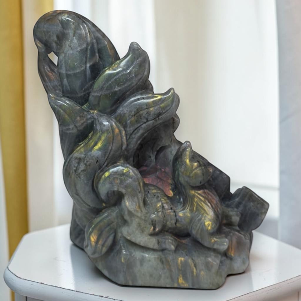 A polished dark gray stone sculpture depicting a fox curled up on a rock, with iridescent gold and green flashes, displayed on a white table against a soft-focus indoor background.
