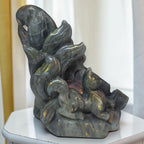 A polished dark gray stone sculpture depicting a fox curled up on a rock, with iridescent gold and green flashes, displayed on a white table against a soft-focus indoor background.