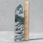Green druzy moss agate tower with white bands beside inch ruler