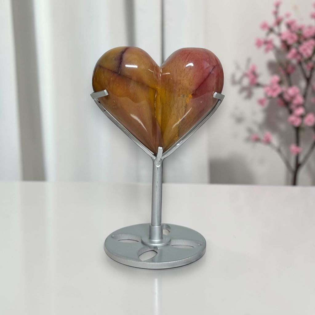 A polished heart-shaped crystal or stone sculpture in warm amber, orange, and reddish tones, displayed on a silver metal stand with a round base on a white tabletop.