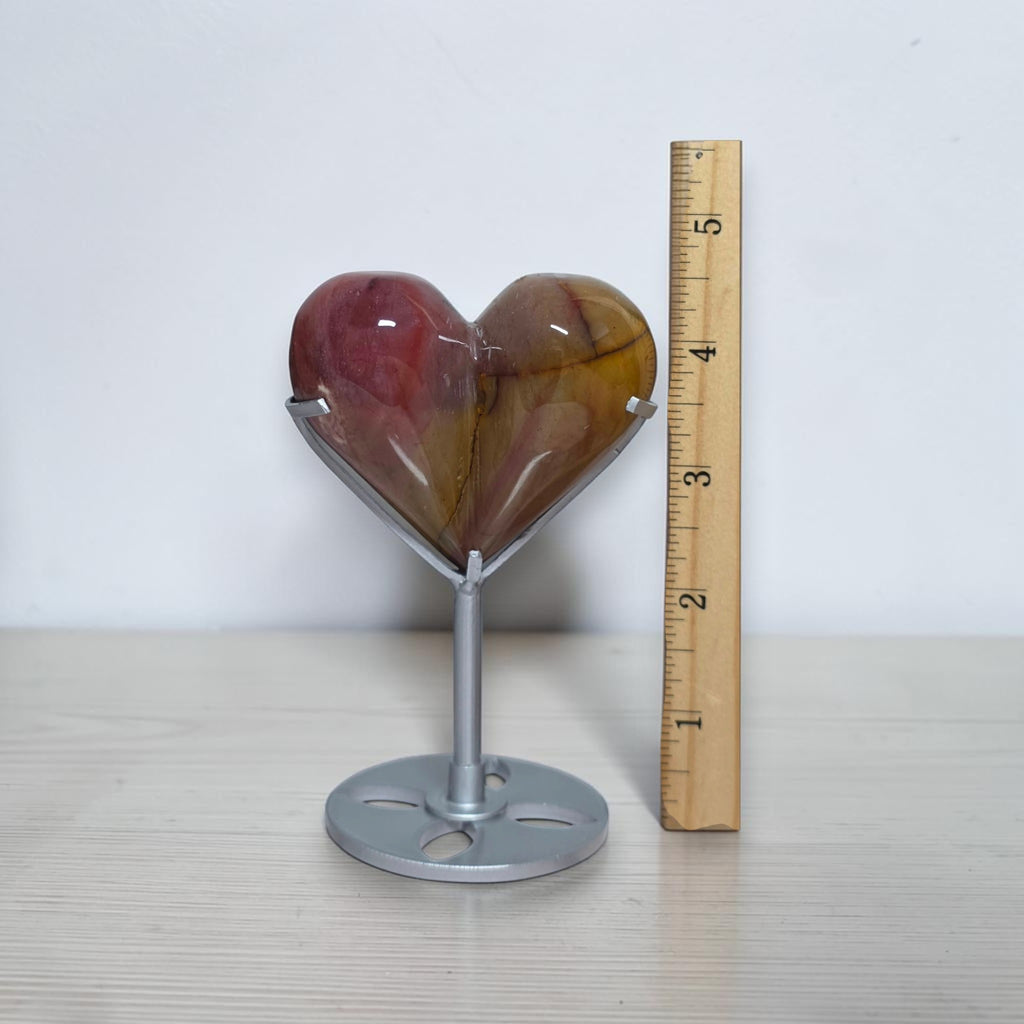 A heart-shaped polished stone sculpture on a silver metal stand, shown next to a wooden ruler for scale on a light wood surface against a white wall.