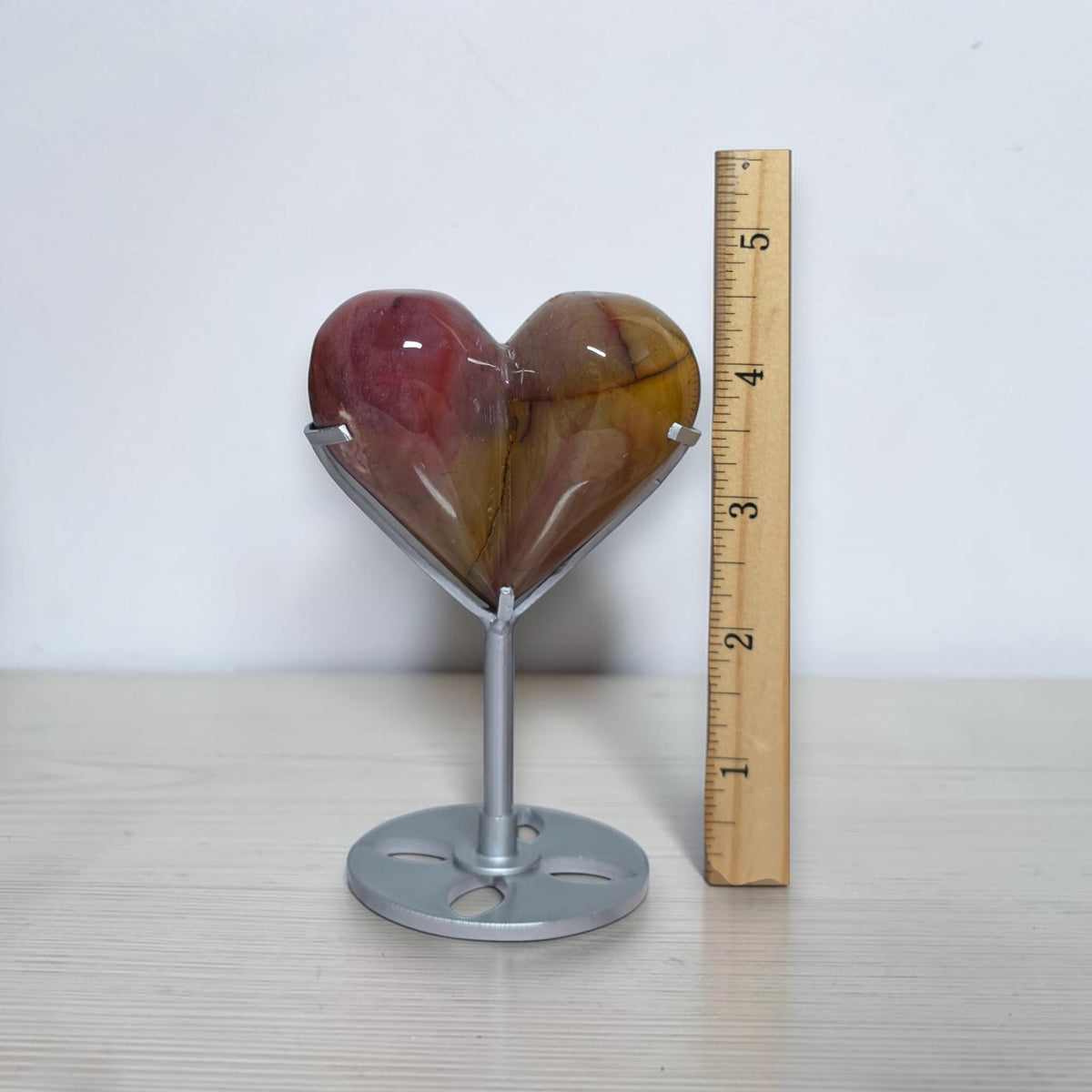 A heart-shaped polished stone sculpture on a silver metal stand, shown next to a wooden ruler for scale on a light wood surface against a white wall.
