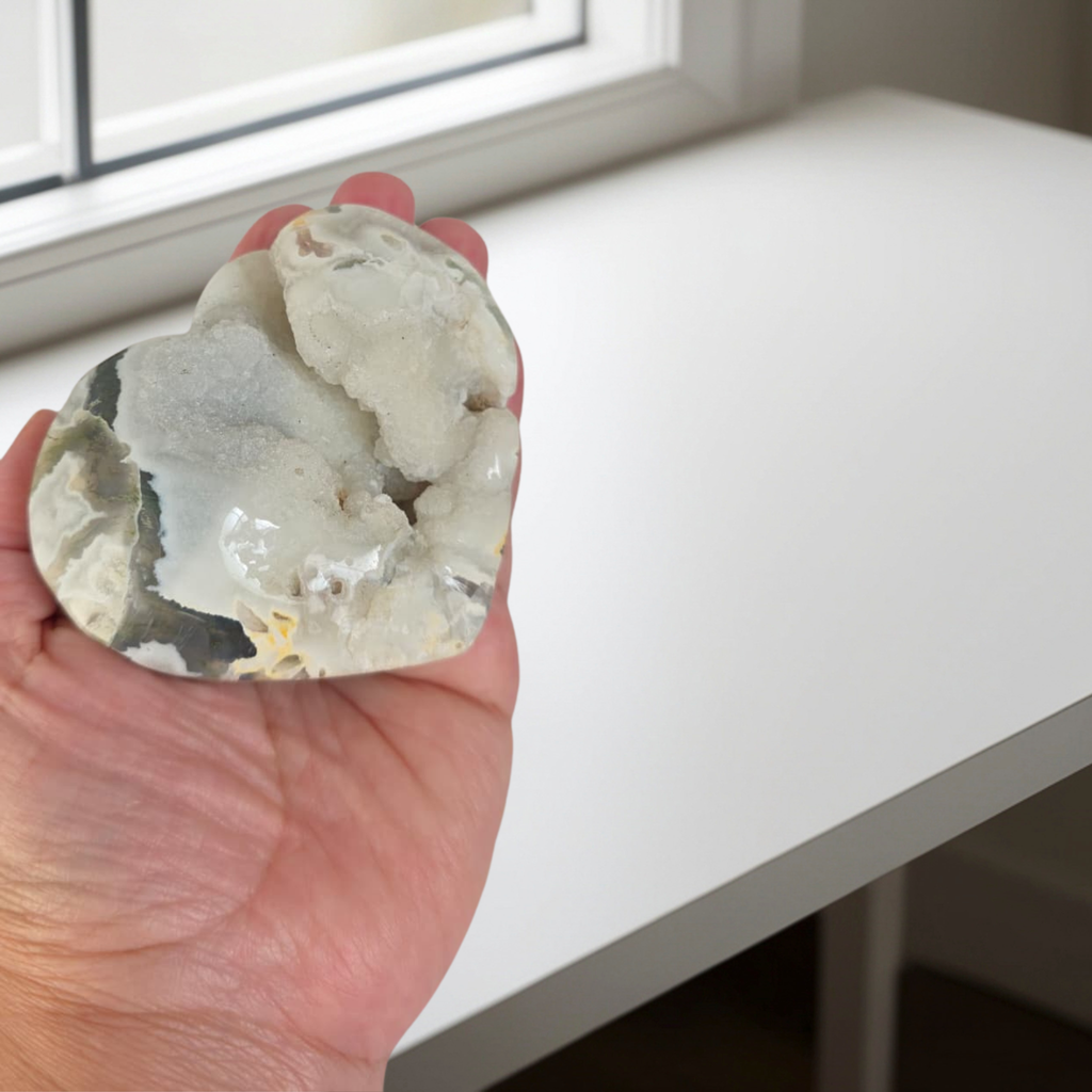 Hand holding a large crystal or mineral stone on a white surface with a window in the background.