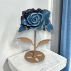 Natural Trolleite rose carving on gold stand displayed by window with blue curtains, front view