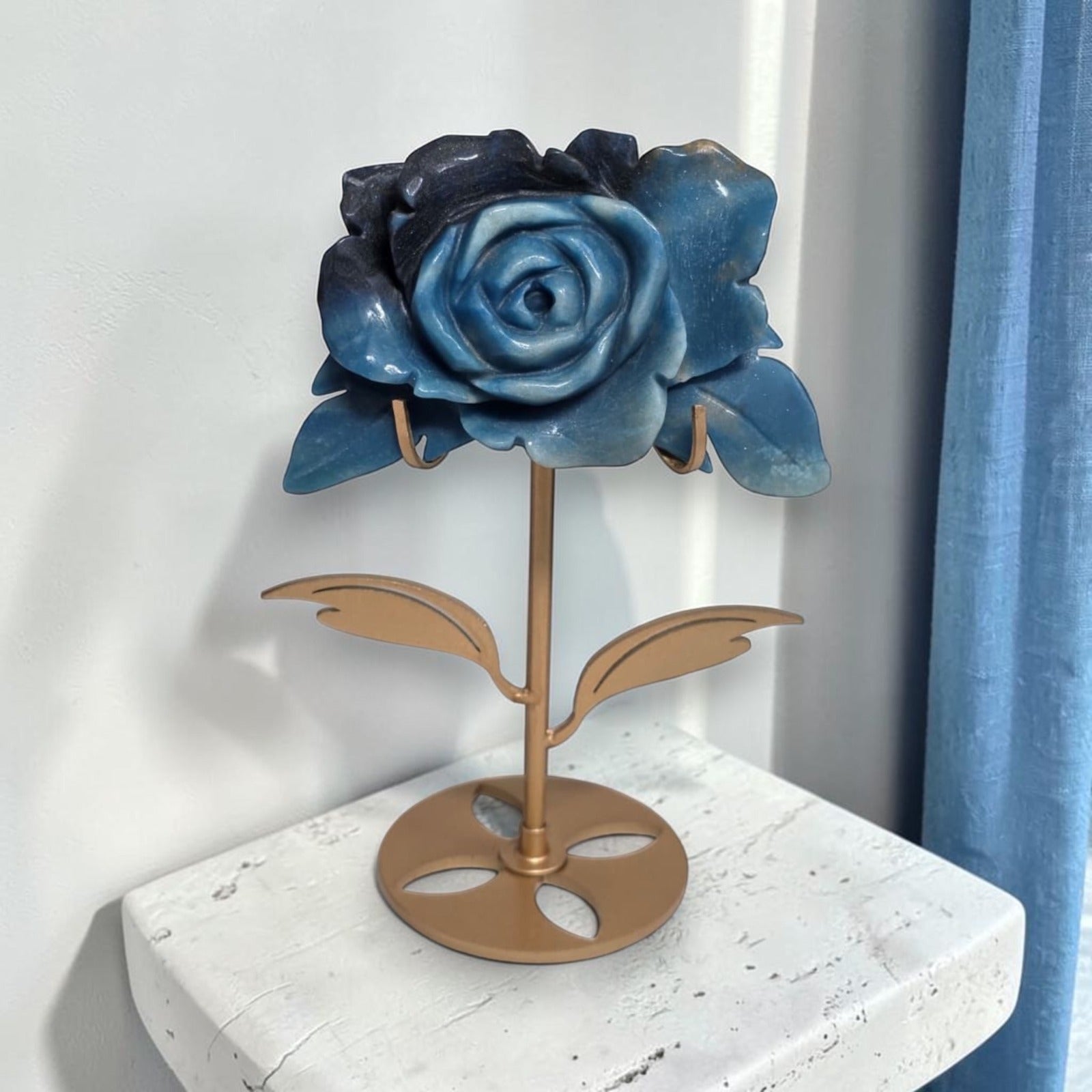Natural Trolleite rose carving on gold stand displayed by window with blue curtains, front view