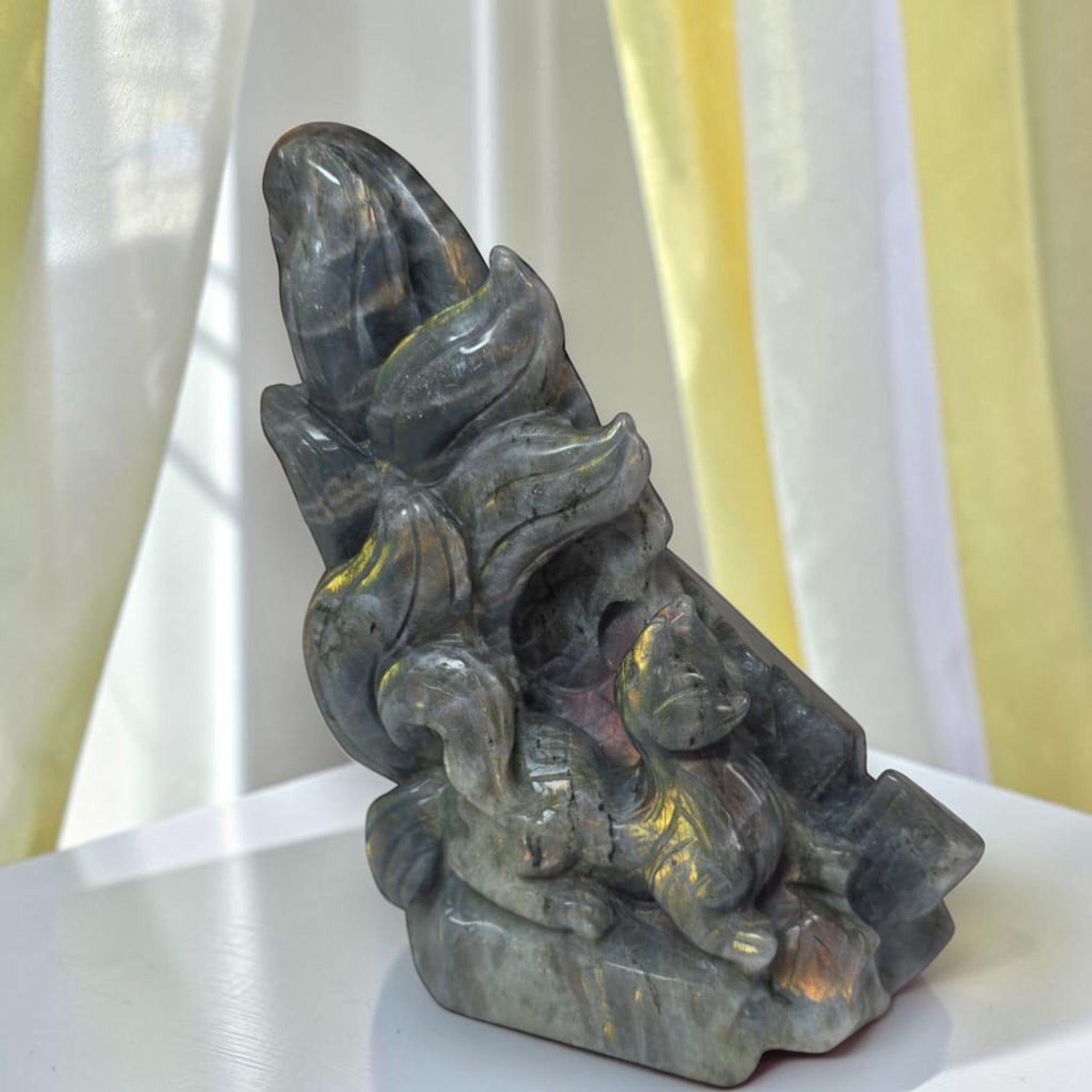 A carved stone figurine of a fox and a small animal, possibly a cat or mouse, set against a background of sheer white curtains with yellow lighting.
