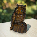 A carved wooden owl figurine with large yellow eyes, perched on a small tree stump base, photographed outdoors on a light surface with a blurred green background.