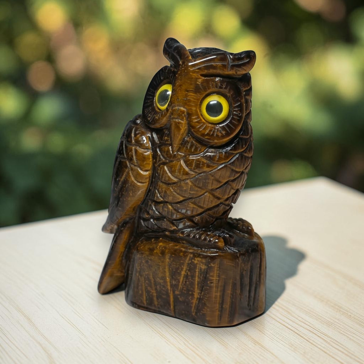 A carved wooden owl figurine with large yellow eyes, perched on a small tree stump base, photographed outdoors on a light surface with a blurred green background.