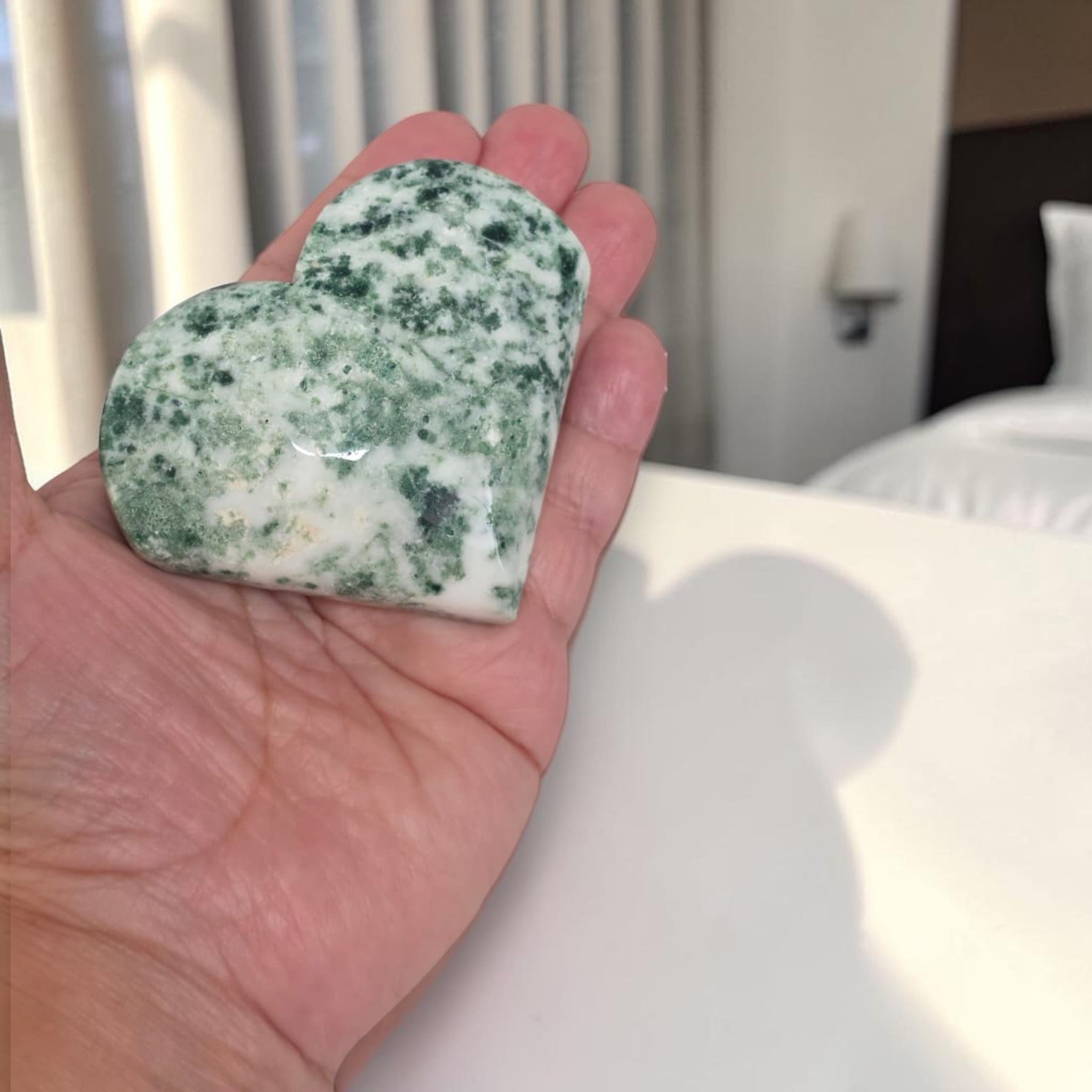 Heart-shaped green and white stone held in a hand with a blurred indoor background